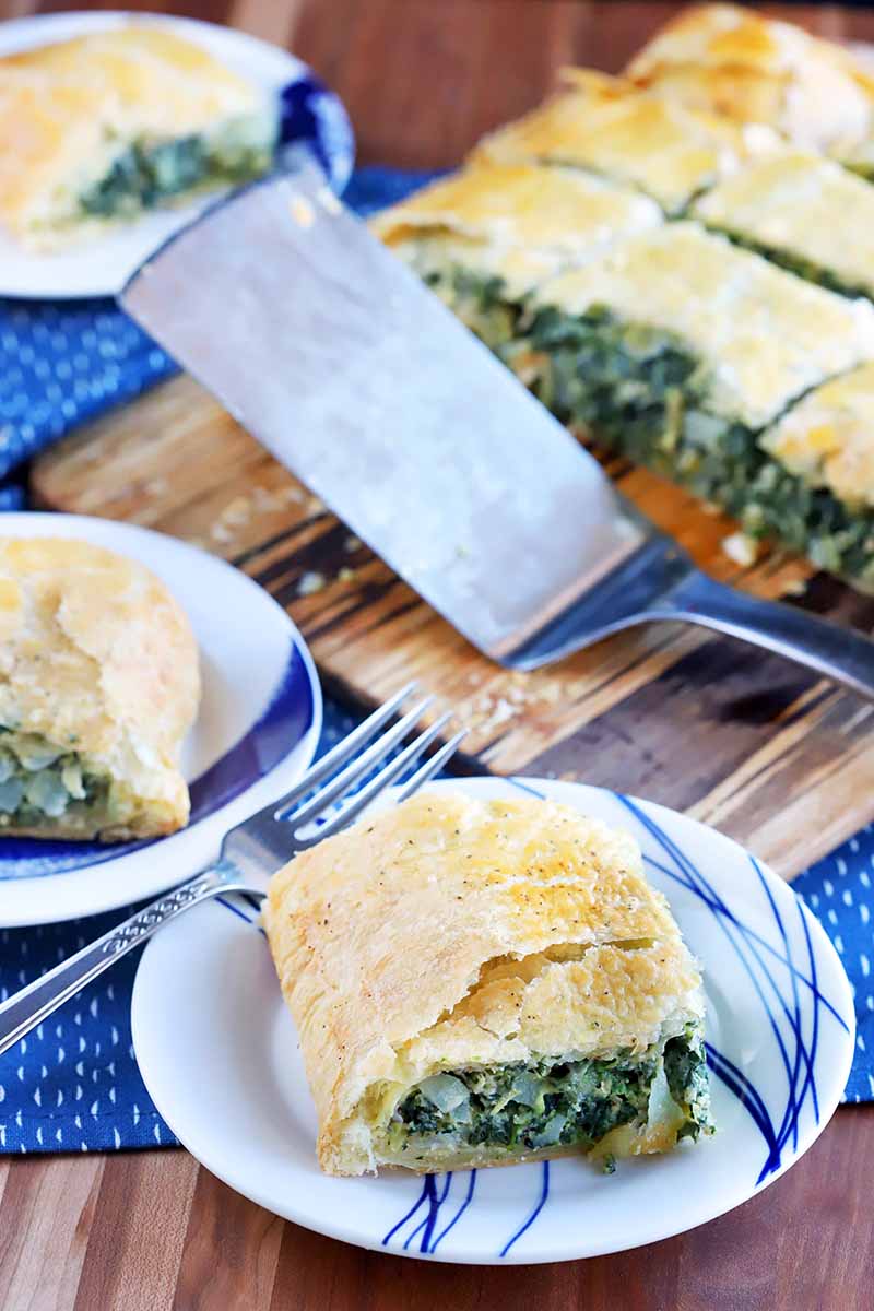 Vertical oblique shot of three plates and a board topped with square portions of spinach in puff pastry, with a metal spatula, on a blue cloth on top of a wood surface.