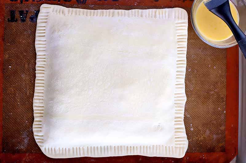 Overhead shot of a square of filled puff pastry with fluted edges on a beige and orange Silpat silicone pan lines, with a small cup of egg wash and a silicone brush.