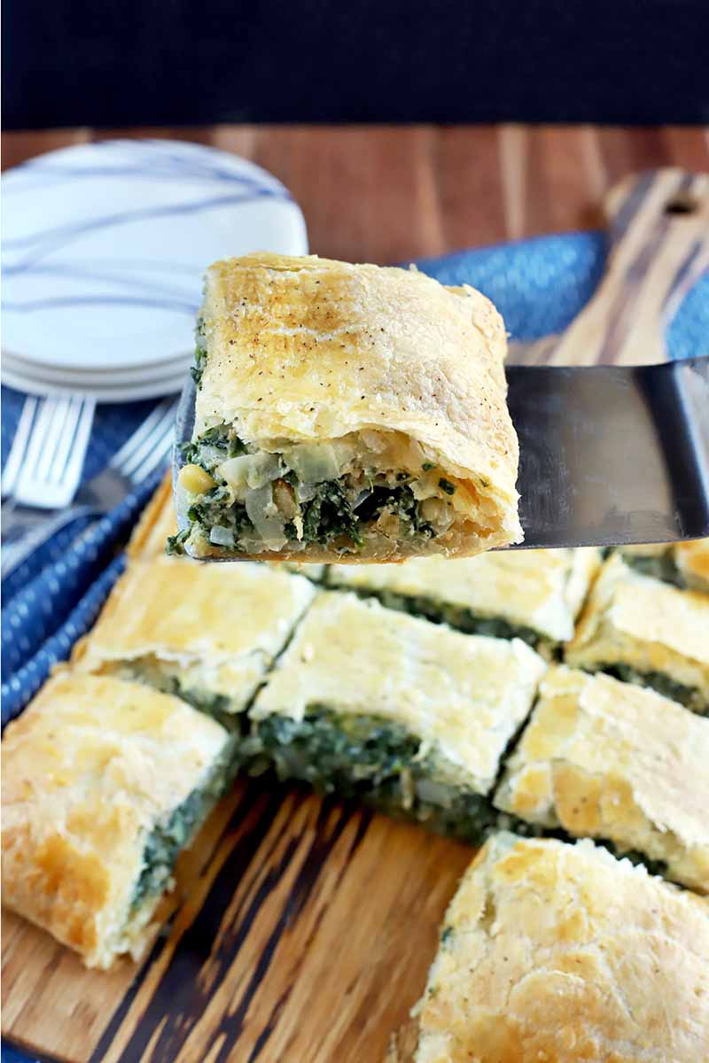 A metal spatula holds a slice of spinach in puff pastry up to the viewer, with more pieces arranged in rows on a wood board in the background, on a blue cloth on top of a wood surface, with a small stack of plates and forks to the left, on a black background.