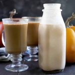 Horizontal image of a bottle of a light orange milky liquid next to pumpkins and glasses of coffee.