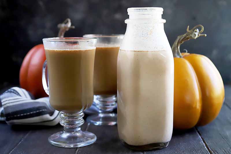 Horizontal image of a bottle of a light orange milky liquid next to pumpkins and glasses of coffee.