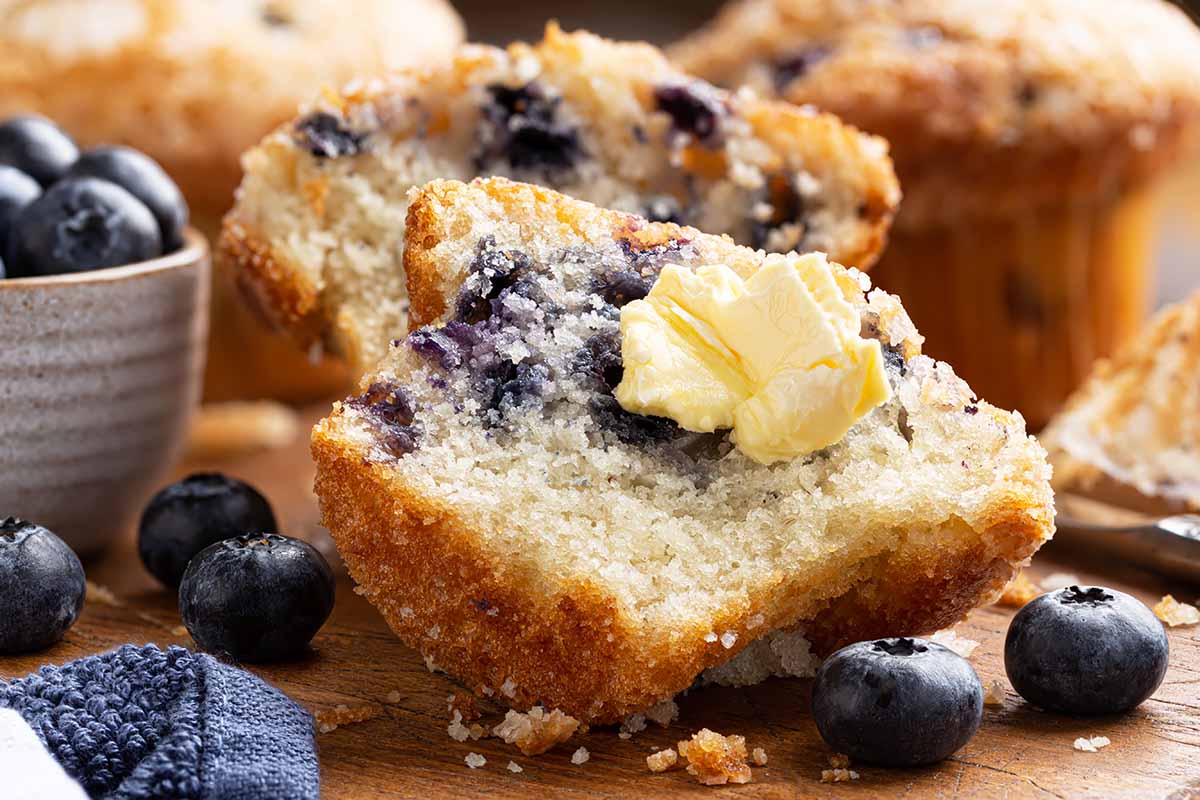 Horizontal closeup image of a halved blueberry pastry with butter spread on one side, with fresh blueberries on a wooden cutting board.