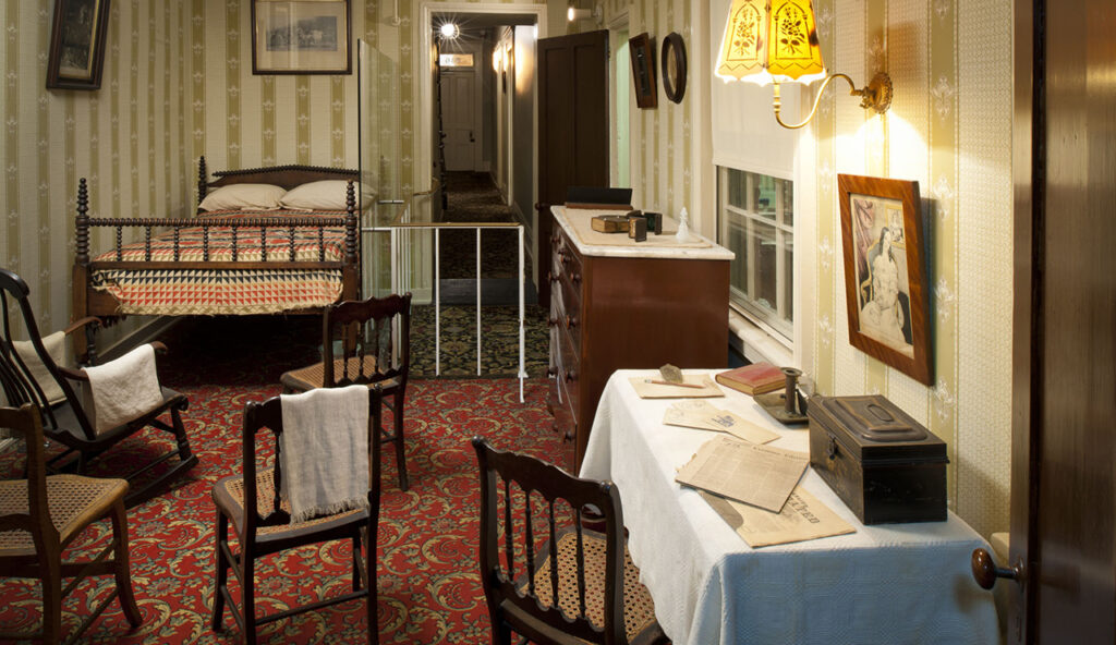 A small bedroom decorated with 1860s-era furniture. The room has green-and-white-striped wallpaper. In the right corner sits a double-sized mattress on a wooden bed frame with two pillows and a colorful quilt with geometric shapes. Small wooden chairs are arranged facing the bed.