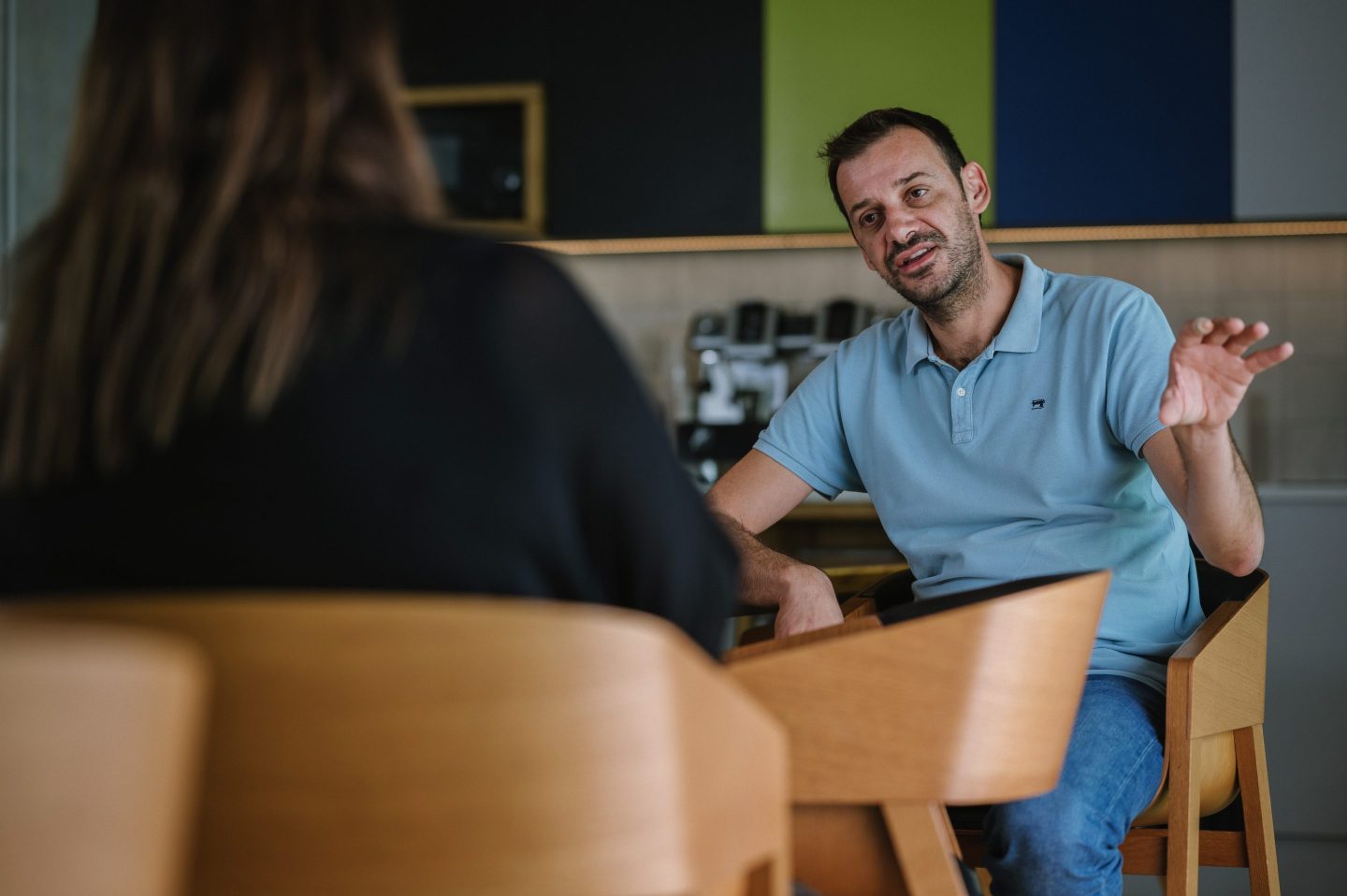 a man seated on a wooden chair, talking to someone facing their back to the camera