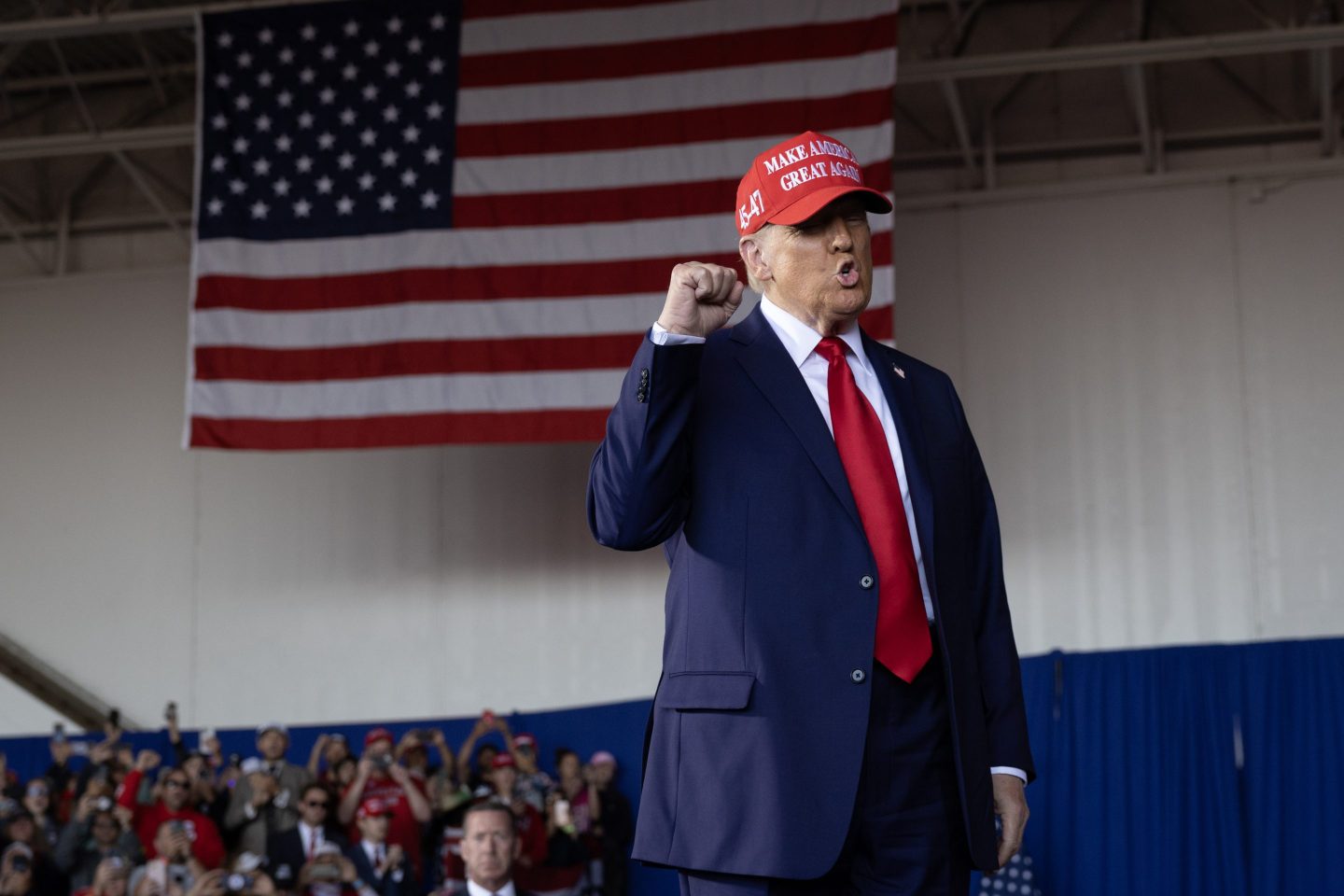 Trump pumps his fist in front of the American flag while wearing a suit and red MAGA hat.