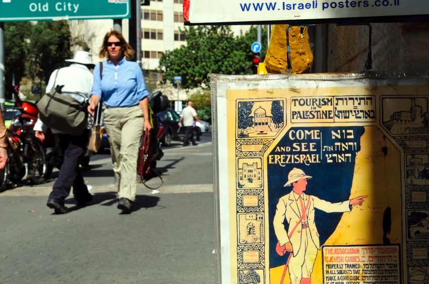 Pedestrians walk past an antique tourism poster for sale outside a shop on a Jerusalem street.  The poster is written in Hebrew and English and uses both the terms Palestine and Eretz Israel.