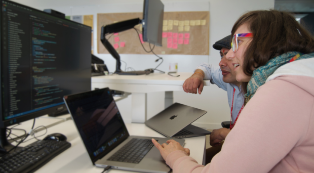 The image features two people sitting at a desk in an office, looking at a computer screen. There are two laptops on the desk in front of them.