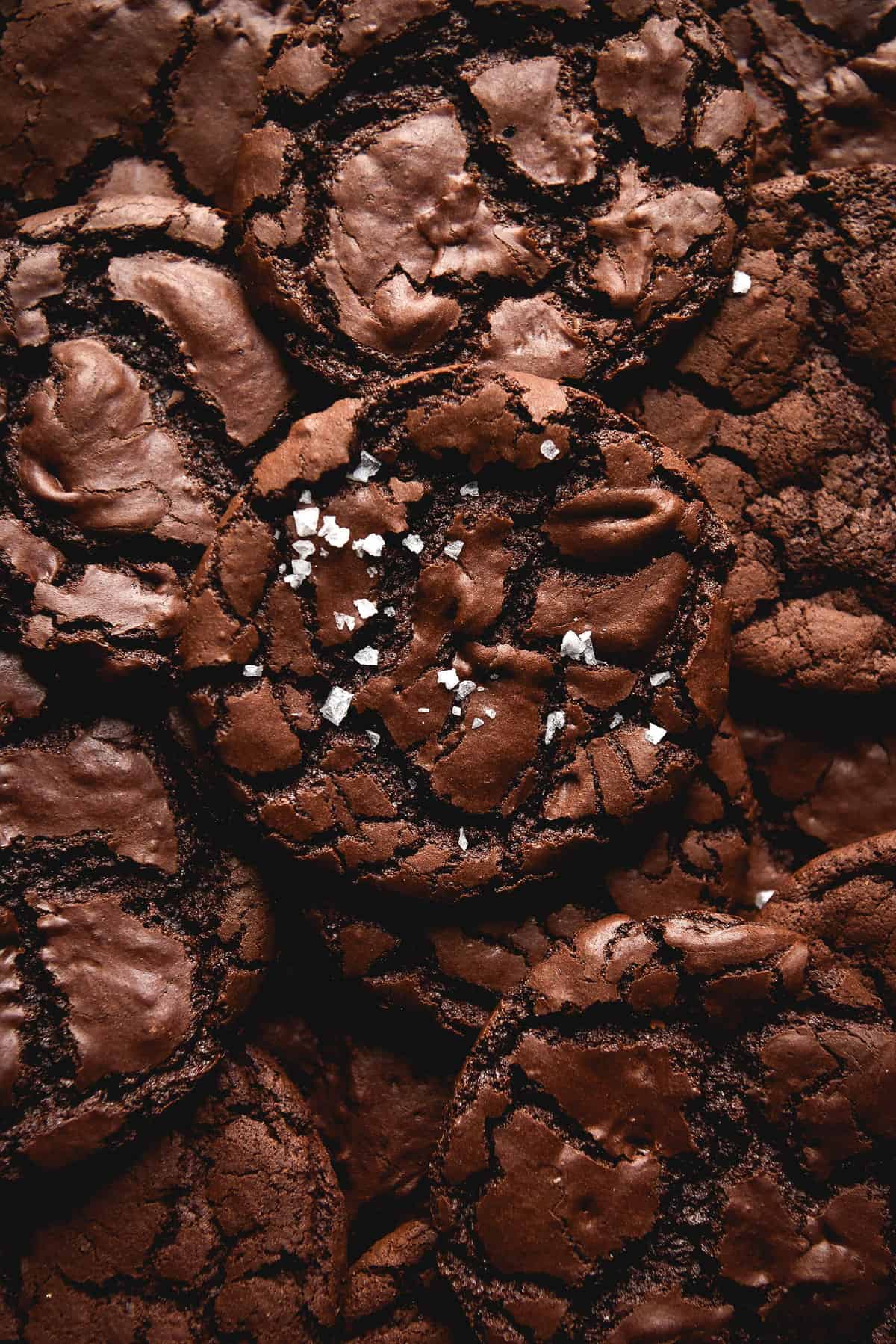 An aerial close up view of a stack of gluten free vegan brownie cookies. The brownie cookies are shiny and crackled and the centre cookie is sprinkled with sea salt flakes