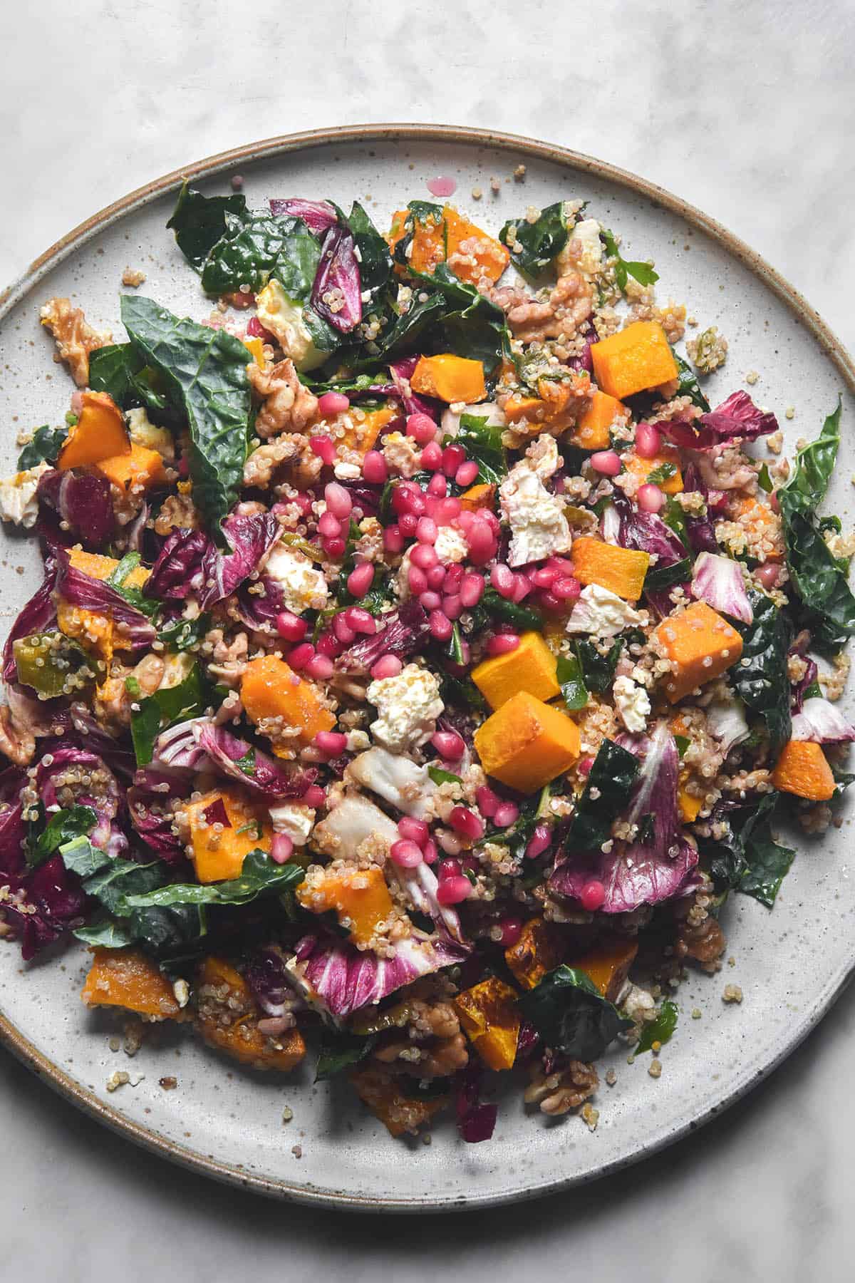 An aerial image of a white serving platter topped with a roast pumpkin, quinoa, feta and kale salad topped with pomegranates and walnuts. The salad sits on a white marble table 