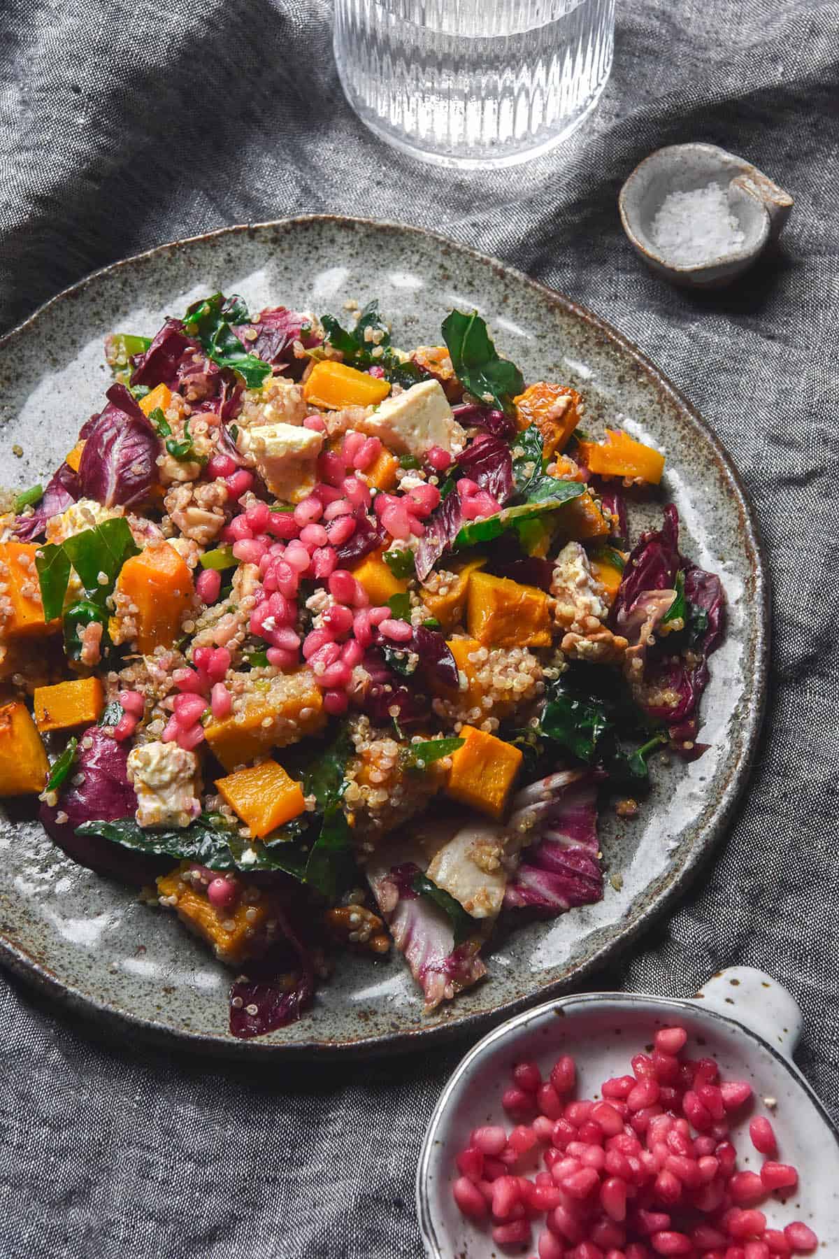 An aerial image of a dark blue ceramic plate topped with a roast pumpkin, quinoa, feta and kale salad topped with pomegranates and walnuts. The salad sits on a dark blue linen tablecloth and is surrounded by a glass of water, a pinch bowl of salt and a white ceramic bowl filled with extra pomegranate arils.