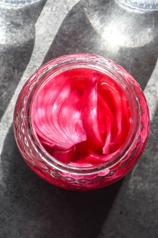 An aerial image of a jar of low FODMAP pickled onions atop a grey backdrop with two sunlit glasses of water in the top of the image. The onions and brine are a vibrant pink purple colour which pops against the grey backdrop.