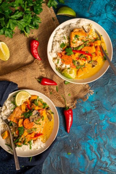 2 bowls of salmon curry over rice with peppers and herbs laying about