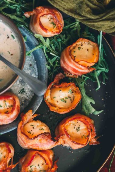 Bacon-wrapped scallops on a black plate with a dipping sauce.