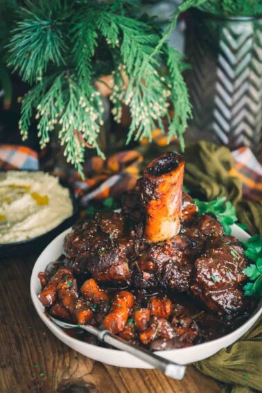 A plate of braised beef shank ribs on a table with a bowl of hummus.