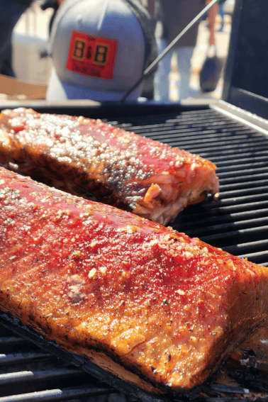 Two large slabs of grilled salmon cooking on a grill with a hat visible in the background.