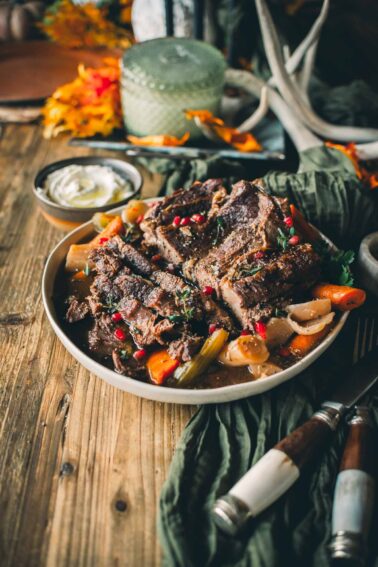 A plate of Instant pot chuck roast with carrots, celery, and pomegranate seeds on a wooden table, surrounded by fall decorations and a small bowl of sauce.