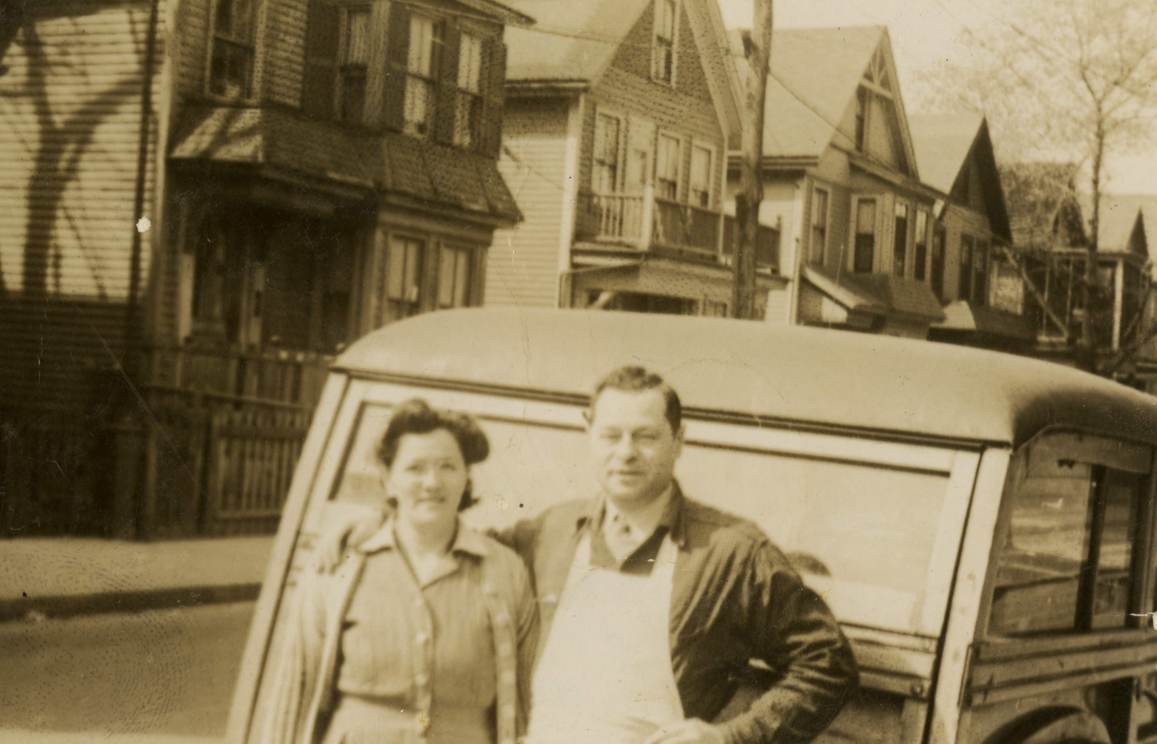 A man and a woman stand in front of a car on a residential street in Dorchester. The man has his hand around the woman's shoulder.