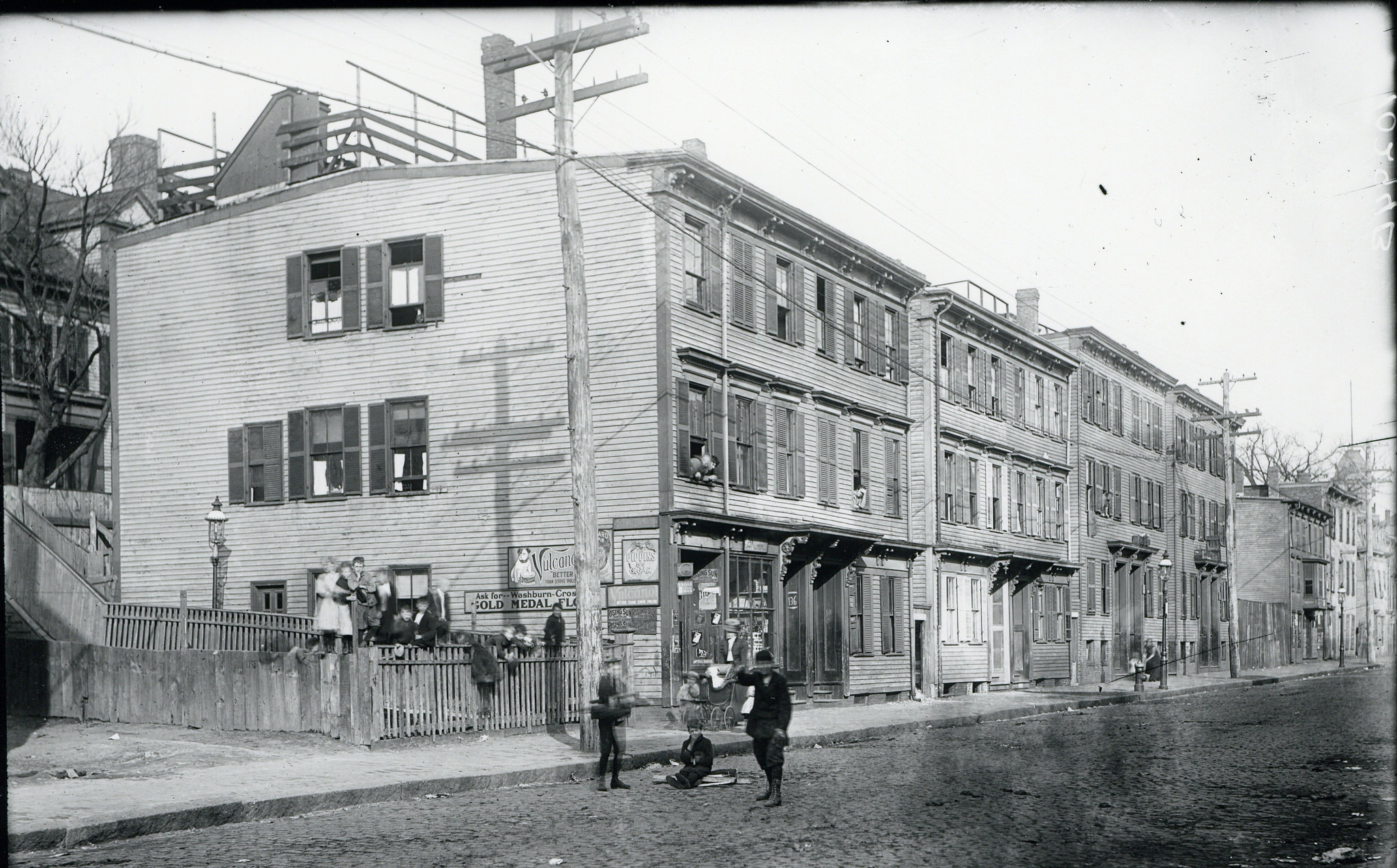 A cobblestoned street with many three story buildings on one side. A group of people gather in front of the first building.