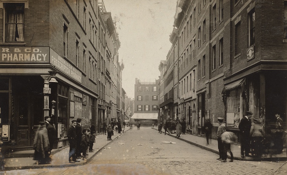 A look down a street in the West End. The road is littered with papers. On one side of the street is a pharmacy. Pedestrians walk along the sidewalk on either side of the street.