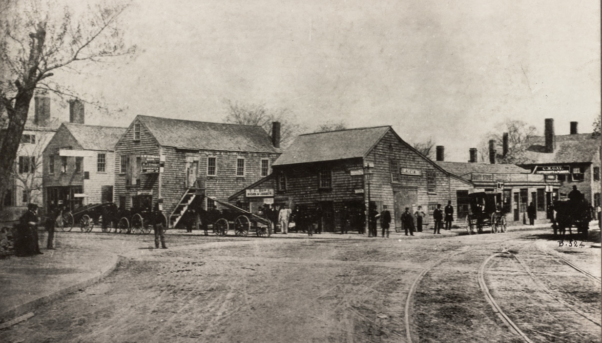 A photograph of an intersection between two dirt roads. Horse-drawn carriages line one of the streets, in front of commercial businesses. The buildings are are all generally two or three stories and are in a colonial style.