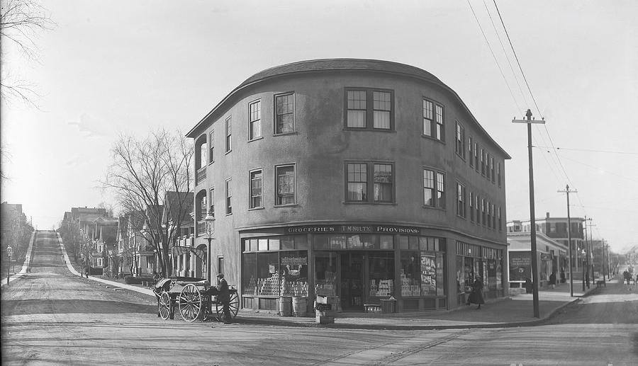 A photograph of Belgrade Avenue and Birch Street. On the corner, in the center of the photograph, is a curved building that is three-stories. On the first floor is a grocer with a sign that reads "T.M. Nulty: Groceries, Provisions." In front of the store is a man with a horse-drawn wagon.