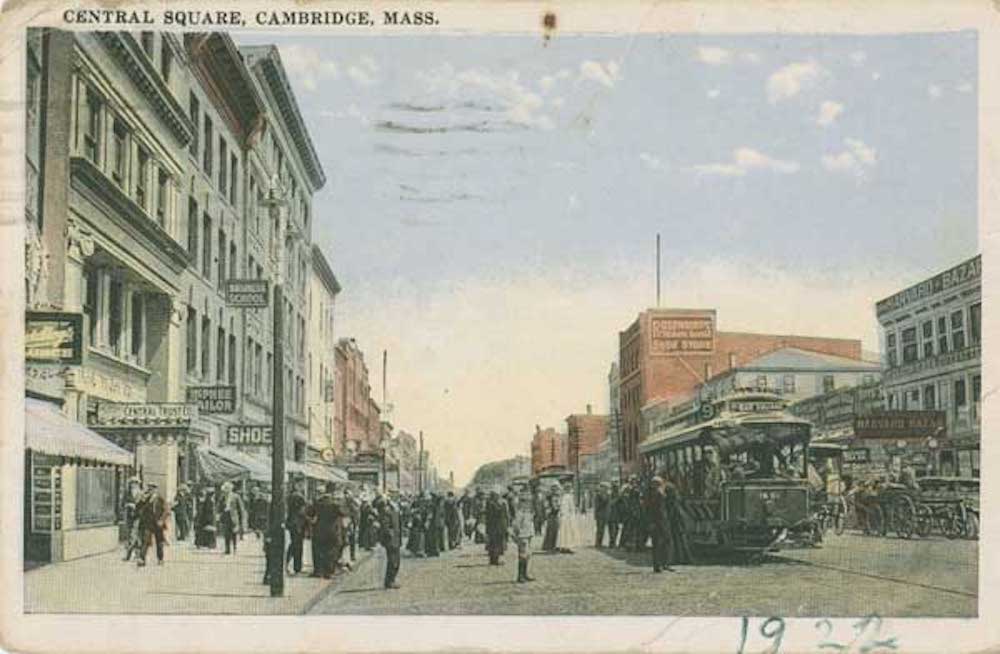 A 1922 postcard of the Central Square in Cambridge Mass. The postcard shows a busy street with many people waiting to board the streetcar in the middle of the street. On either side of the street are businesses and individuals walking on the sidewalks.