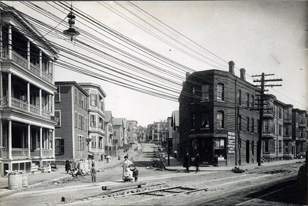 A photograph of an intersection with a streetcar line and a woman and two children in the foreground. In the background, there is a residential neighborhood with three-decker homes.