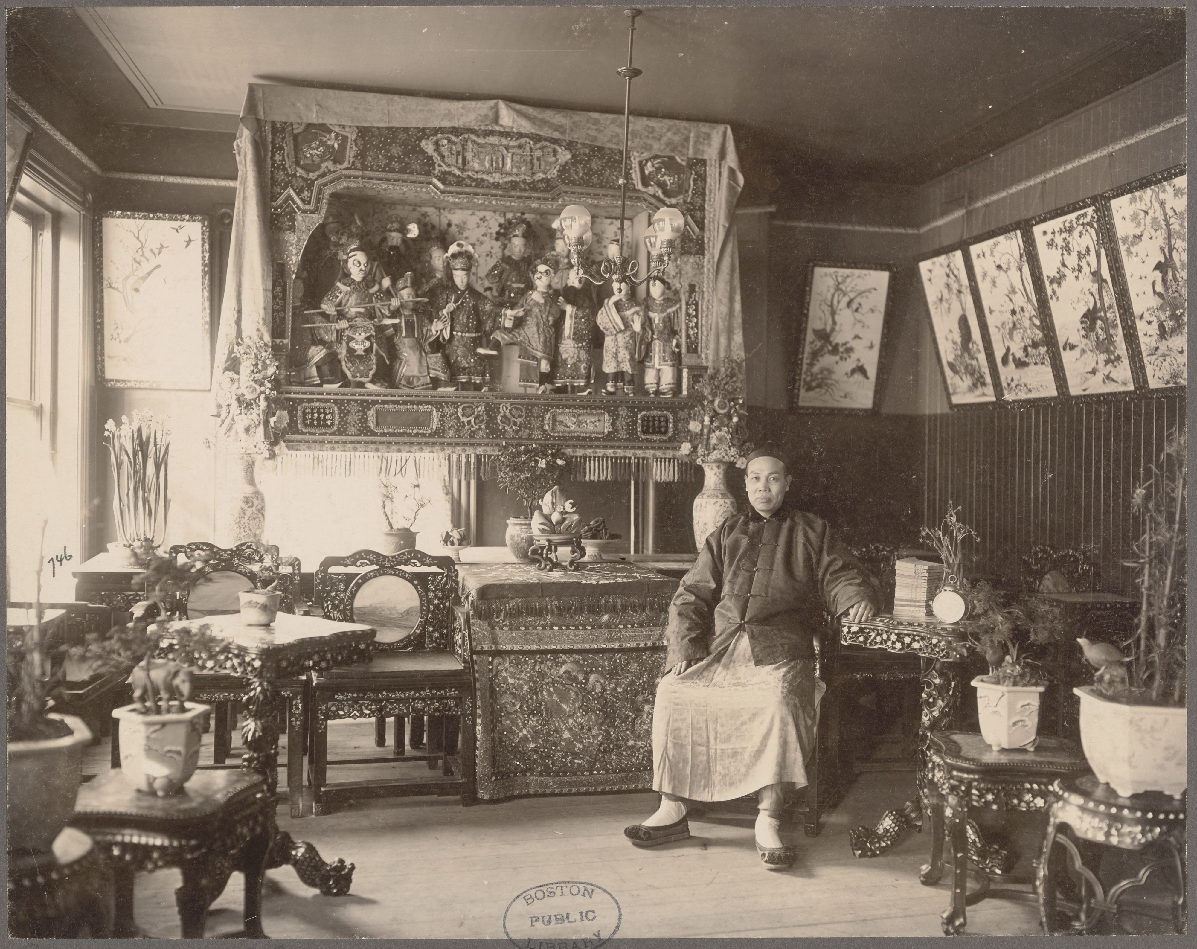 A man in Chinese dress sits in a Chinese restaurant with Chinese style furnishings, wall hangings, and plants..