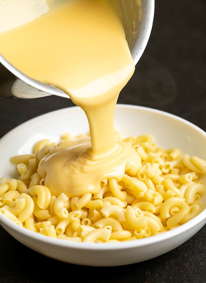 Queso style cheese sauce being poured from a small saucepan on top of a white bowl of cooked elbow macaroni