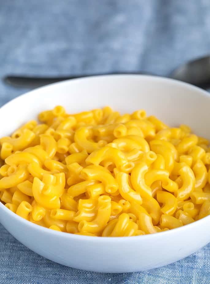 gluten free mac and cheese in white bowl with bowl of cooked elbows with powdered cheese-base cheese sauce and spoon in background on blue cloth