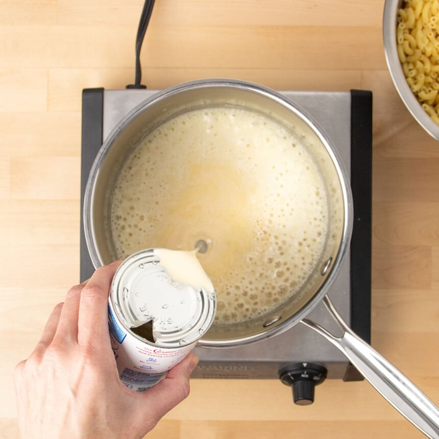 hand pouring evaporated milk from a metal can into a metal saucepan with light tan bubbling sauce