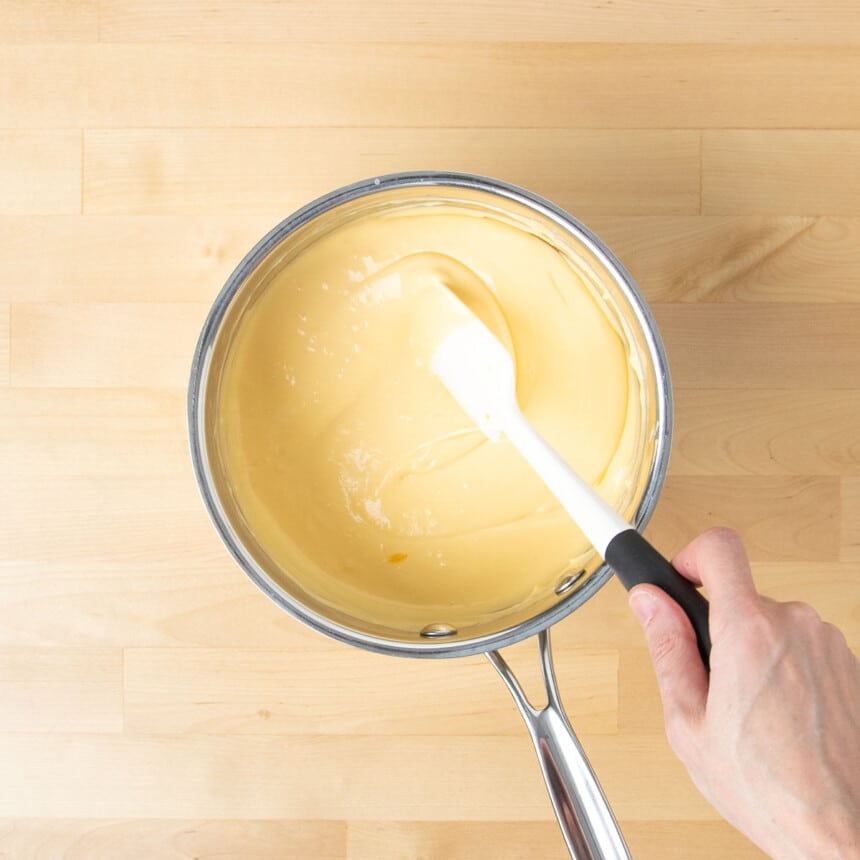 hand holding white silicone spatula mixing and turning over light yellow cheese sauce in saucepan