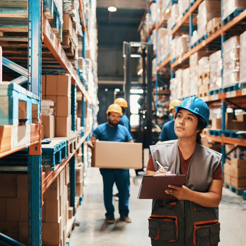 Young woman wearing turquoise hard hat holding clipboard and walking in warehouse