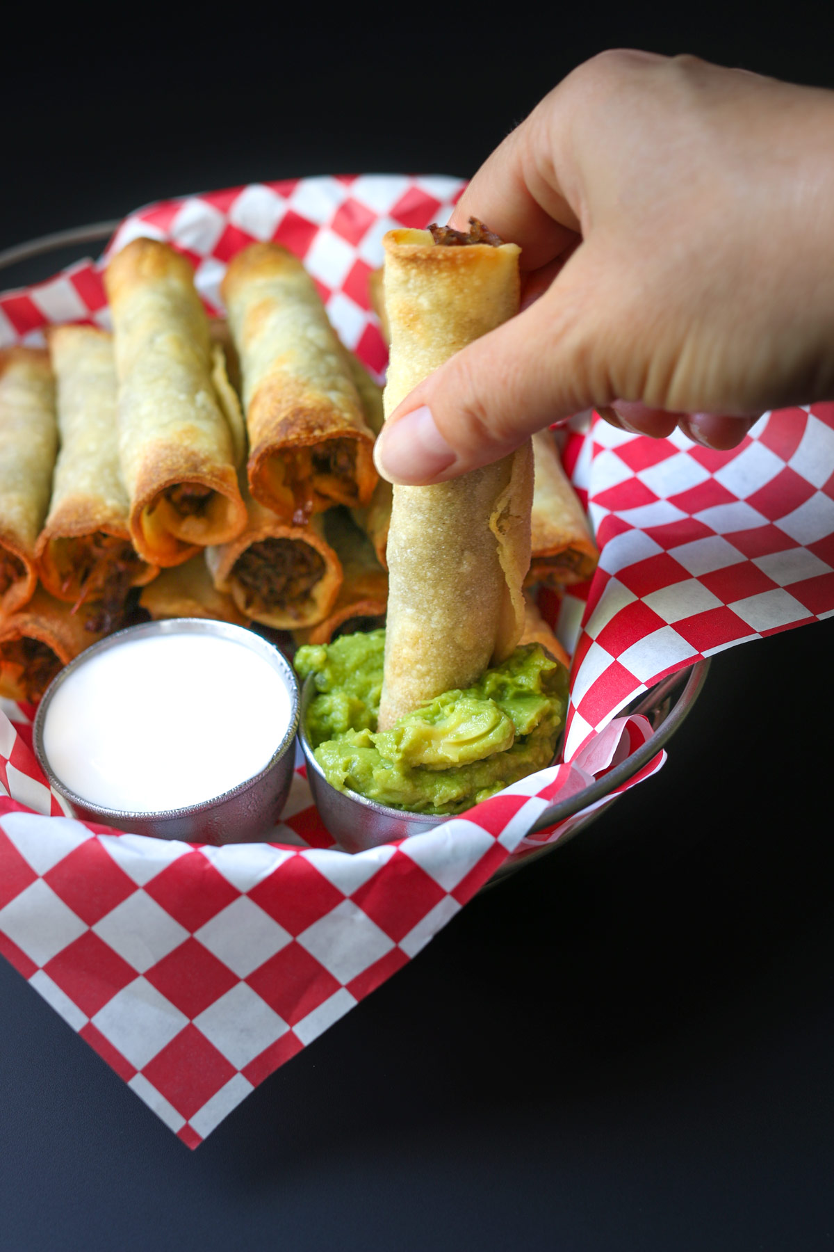 hand dipping beef taquito into guacamole next to sour cream cup and more taquitos.