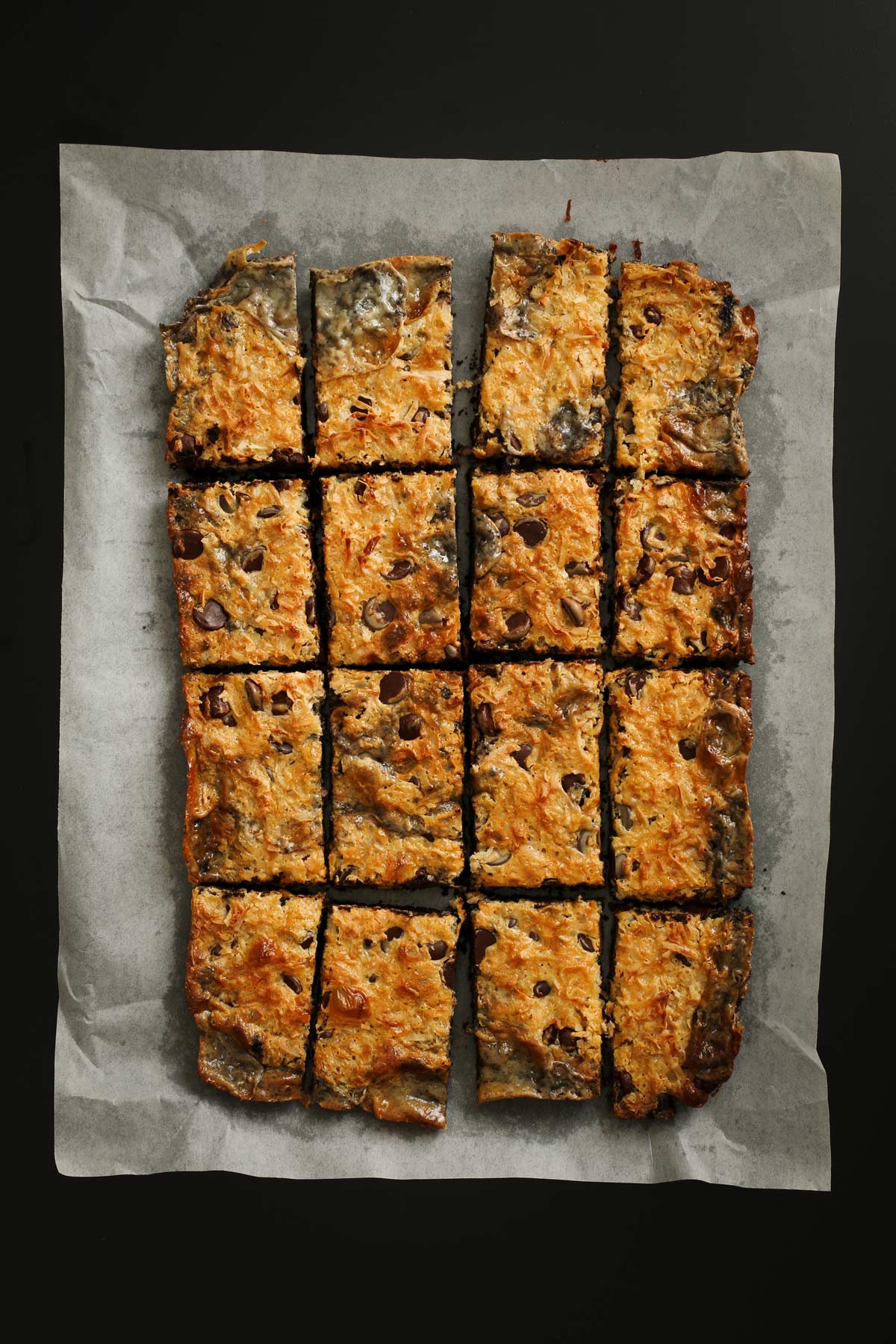 flat of chocolate coconut bars cut into slices on parchment paper on a black table.