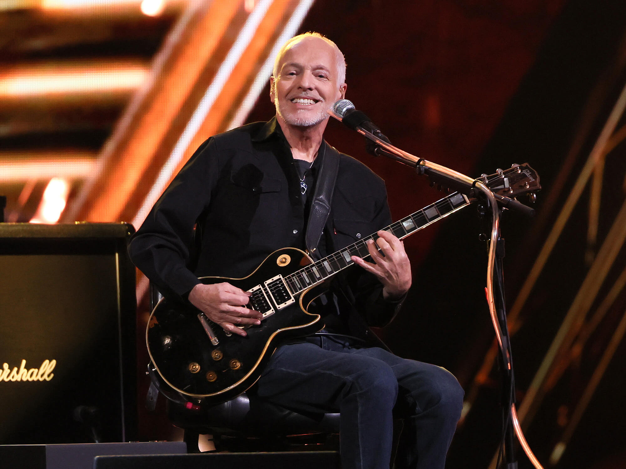 Peter Frampton on stage. He is sitting down and playing a black Les Paul with a smile on his face.