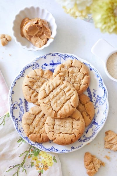 A plate of peanut butter cookies arranged on a decorative blue and white plate. A small bowl of peanut butter, a jug with brown sugar, and cookie crumbs are beside the plate. A pink and green floral towel and yellow flowers partially frame the scene.