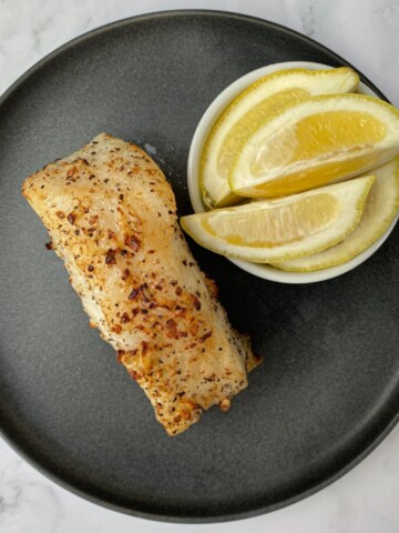 Air fried Chilean sea bass fillet on a plate with lemon wedges.