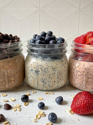 Three mason jars filled with protein overnight oats. The three flavors include: Chocolate chip overnight oats have chocolate chips on top, blueberry cheesecake overnight oats has blueberries on tope, and strawberry shortcake overnight oats has chopped strawberries on top.