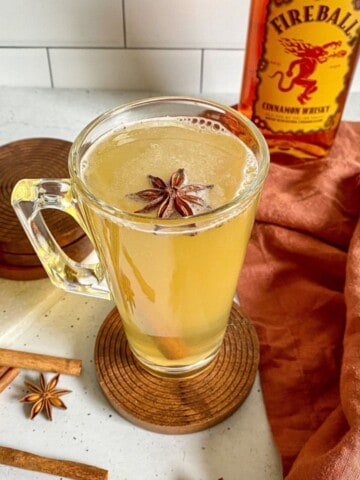 Hot Toddy in a clear mug surrounded by sliced lemon and cinnamon sticks. A bottle of Fireball whisky is next to the cocktail.