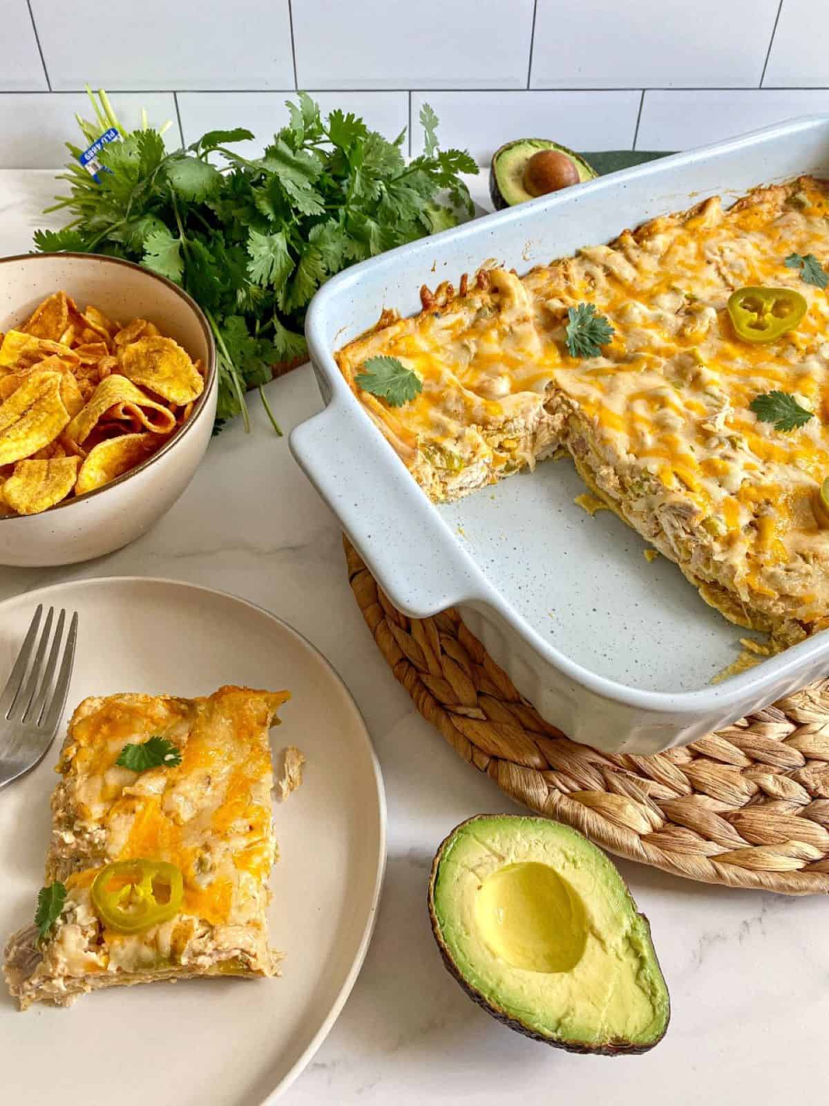 Sour Cream Chicken Enchilada Casserole cut into with one serving of the layered chicken casserole on a plate. Cilantro, avocado, and chips surround the meal.