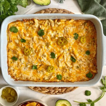 Sour Cream Chicken Enchilada Casserole in a large baking dish. A green kitchen towel, cilantro, avocado, and chips are around the dish.