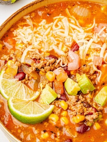 Beef enchilada soup made with ground beef and red enchilada sauce in a bowl with corn, black beans, cheese, avocado slices, and lime wedge.