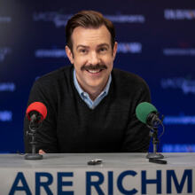 A smiling man with a mustache sits behind two colorful microphones.