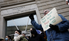 A protester stands outside the USAID building holding up a sign that reads, "Musk and Trump: keep your fascist hands off USAID"