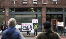 A groups stands outside a Tesla showroom holding signs, one of which says "Tesla funds fascists."