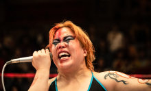 Professional wrestler with dramatic face paint and a microphone, passionately speaking during a match.