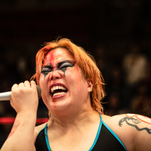 Professional wrestler with dramatic face paint and a microphone, passionately speaking during a match.