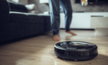 A robot vacuum on a wooden floor. A person's legs are seen running behind it.