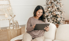 a person sits on a white couch while looking at a tablet and smiling. In the background, a lit up holiday tree